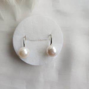 May include: A pair of pearl drop earrings. Each earring has a lustrous, round, off-white pearl suspended from a silver-coloured hook. The earrings are displayed on a white, circular backing against a soft, white background.