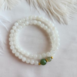 May include: A white beaded bracelet with a green and gold accent. The bracelet is composed of small, round, white beads. The accent includes a larger green bead and a smaller, lighter green bead, connected by gold-coloured metal.