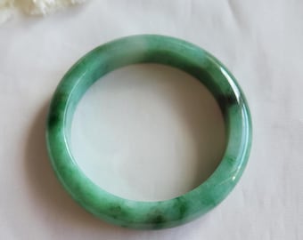 Op de afbeelding: Een ronde jade armband. De armband heeft een levendige groene kleur met donkerdere groene en witte adering. Het gladde, gepolijste oppervlak reflecteert licht en benadrukt de natuurlijke variaties in de steen. Een klassiek sieraad.