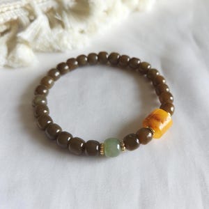 Könnte beinhalten: Ein Armband mit Perlen mit dunkelbraunen, abgerundeten Perlen. Das Armband enthält eine hellgrüne Perle und eine rechteckige, bernsteinfarbene Perle. Goldfarbene Akzente sind zwischen einigen Perlen sichtbar. Das Armband wird auf einer weißen Oberfläche präsentiert.