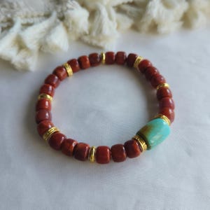 Könnte beinhalten: Ein Perlenarmband mit runden, rotbraunen Steinperlen, die mit kleinen, polierten goldfarbenen Metallzwischenstücken durchsetzt sind. Eine einzelne, größere zylindrische türkisfarbene Perle befindet sich an einer Seite, akzentuiert durch goldfarbene Metallringe.