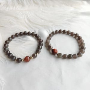 May include: Two beaded bracelets. Each bracelet features dark gray beads with a reddish-brown accent bead. The bracelets are on a white surface. The beads are strung on an elastic cord.