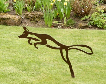 Puede incluir: Una silueta de metal de un zorro saltando en un jardín de hierba. El zorro es marrón y se recorta contra un césped verde.
