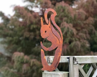 Puede incluir: Una estaca de jardín con silueta de ardilla de metal oxidado. La ardilla está representada en una pose estilizada, con la cola curvada hacia arriba. La estaca de jardín está montada en un poste de madera, con un fondo borroso de follaje verde y marrón.