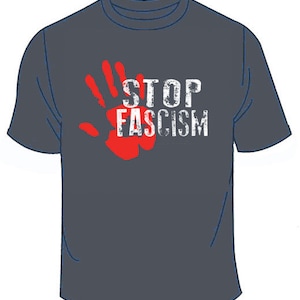 Puede incluir: Camiseta gris oscuro con una huella de mano roja y las palabras "STOP FASCISM" en letras blancas desgastadas. La camiseta tiene cuello redondo y mangas cortas. El diseño está centrado en la parte delantera.