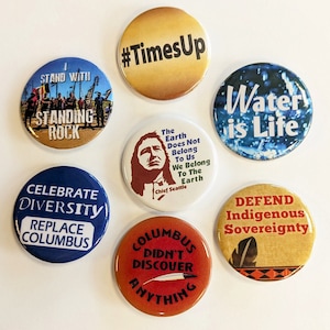 May include: Nine round buttons with various messages and designs. The buttons are colorful and feature messages such as "Stand with Standing Rock", "Celebrate Diversity", "Replace Columbus", "#TimesUp", "The Earth Does Not Belong To Us", "Water is Life", "Columbus Didn't Discover Anything", and "Defend Indigenous Sovereignty".