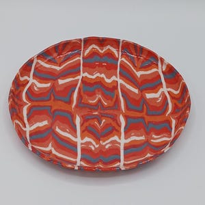May include: Oval ceramic serving dish with a vibrant, abstract pattern. The dish features a base of orange and red hues, overlaid with wavy lines in white and blue. The design creates a dynamic, eye-catching aesthetic.