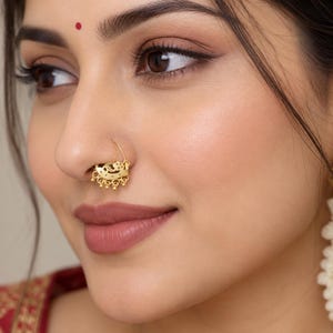 May include: A gold nose ring featuring a crescent moon shape with intricate cut-out patterns and small dangling bead accents. The jewellery piece is worn on the side of the nostril, showcasing a polished metallic finish and detailed filigree work.