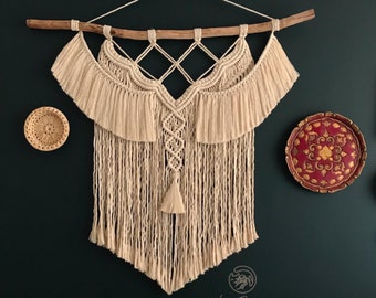 May include: A beige macrame wall hanging with a wooden dowel, featuring a geometric design and fringe. The wall decor is displayed against a dark green wall, with decorative plates and pillows in the background.
