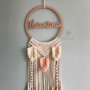 May include: A macrame wall hanging with a wooden ring that says "Valentina" in cursive. The macrame is made with natural coloured cord and has pink and white feathers and tassels.