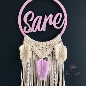 May include: A pink wooden hoop with the name "Sare" written in cursive. The hoop is attached to a macrame dream catcher with white and pink feathers and tassels.