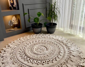 May include: A large, round, beige crocheted rug with a detailed mandala pattern in the centre. The rug is on a wooden floor in a room with a bookshelf and plants.