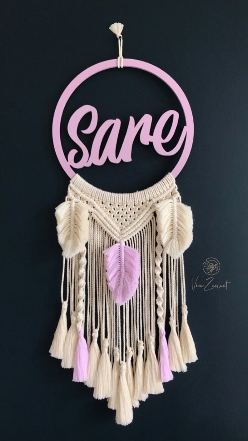 May include: A pink wooden hoop with the name "Sare" written in cursive. The hoop is attached to a macrame dream catcher with white and pink feathers and tassels.