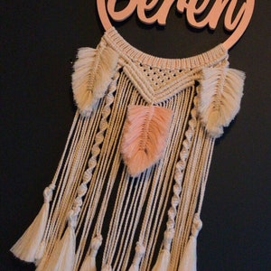 May include: A macrame dream catcher with a wooden hoop that says "Beren" in cursive. The dream catcher has a white and cream colour scheme with peach coloured feathers.