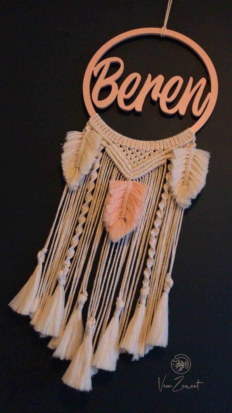 May include: A macrame dream catcher with a wooden hoop that says "Beren" in cursive. The dream catcher has a white and cream colour scheme with peach coloured feathers.