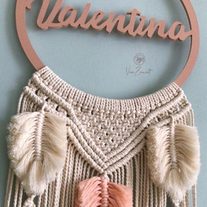 May include: A macrame wall hanging with a wooden ring featuring the name "Valentina". The hanging has a cream-coloured macrame design with three white and peach feathers.