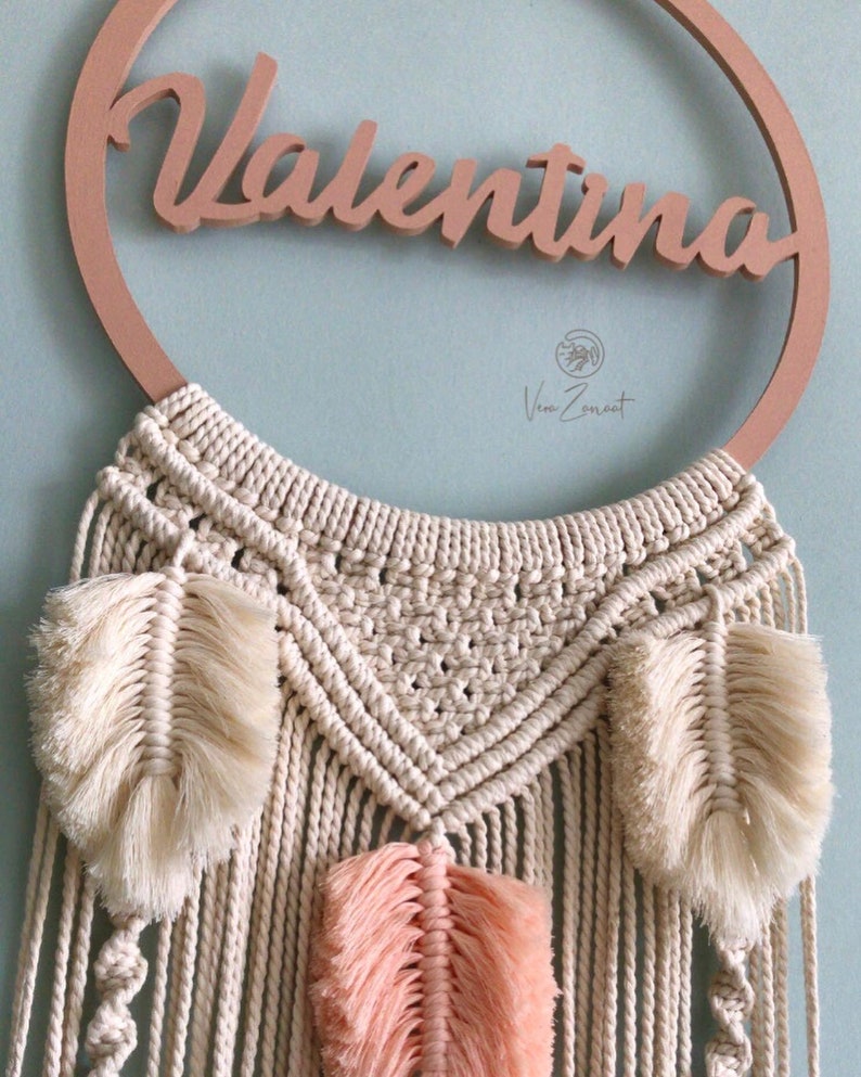 May include: A macrame wall hanging with a wooden ring featuring the name "Valentina". The hanging has a cream-coloured macrame design with three white and peach feathers.