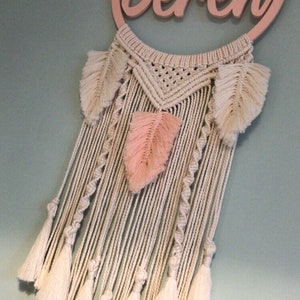 May include: A pink wooden hoop with the name "Beren" written on it. The hoop is attached to a macrame wall hanging with white and pink feathers and tassels.