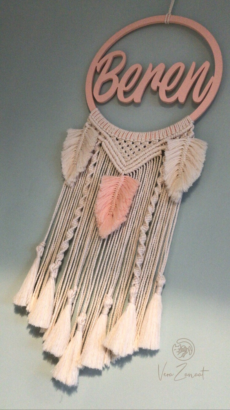 May include: A pink wooden hoop with the name "Beren" written on it. The hoop is attached to a macrame wall hanging with white and pink feathers and tassels.