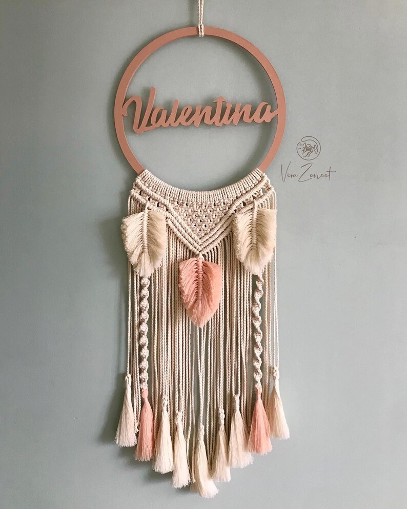 May include: A macrame wall hanging with a wooden ring that says "Valentina" in cursive. The macrame is made with natural coloured cord and has pink and white feathers and tassels.