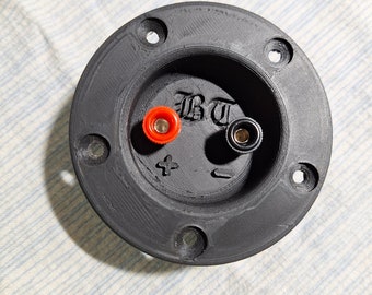 May include: A circular black speaker terminal cup featuring four mounting holes, a red positive binding post, and a black negative binding post. The center of the cup is embossed with the letters BT, a plus sign, and a minus sign symbol.