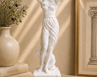 May include: A white marble-finish statue of a standing figure with long wavy hair, hands raised behind the head, wearing a draped garment. The rectangular base features the Greek text ΑΦΡΟΔΙΤΗ. The sculpture is positioned on a flat surface.