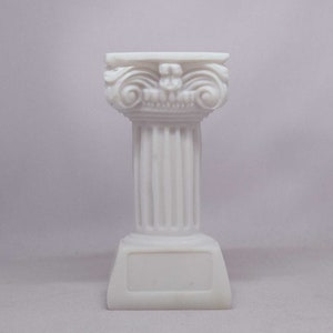 May include: A white, miniature Ionic column with detailed carvings. The column features a fluted shaft, a decorative capital with scroll-like volutes, and a rectangular base. The overall design is reminiscent of classical architecture.