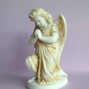 May include: A white marble statue of a kneeling angel with wings. The angel is wearing a flowing robe and has its hands clasped in prayer. The statue has gold accents.