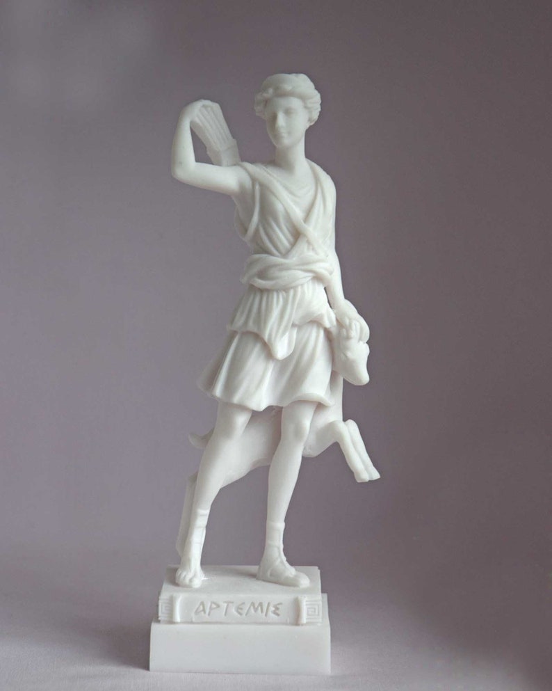 May include: White marble statue of Artemis, the Greek goddess of the hunt, with a bow and arrow and a deer at her side. The statue is on a white marble base with the inscription "ARTEMIS" in Greek.