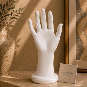 Può includere: Un espositore per gioielli a forma di mano bianca con palmo aperto e dita rivolte verso l'alto. La scultura presenta una finitura liscia e opaca e poggia su una base circolare. Un piccolo cartoncino quadrato con il testo "estatueshop Greek statues" è posizionato accanto alla base.