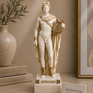 May include: A white and gold statue of the Greek god Apollo standing on a rectangular base with the Greek text ΑΠΟΛΛΩΝ inscribed on the front. The figure wears a draped cloth, gold arm cuffs, and gold sandals while holding a small golden lyre.