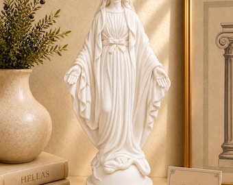 May include: A white statue of the Virgin Mary standing on a globe with a serpent at her feet. The figure wears a draped robe and veil with arms outstretched. A small card displays the text "estatueshop greek statues" in black font.