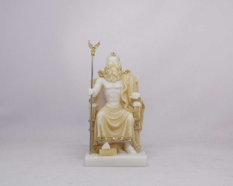 May include: A cream-colored statue of a seated figure, possibly Zeus, holding a staff with a golden eagle at the top. The figure is draped in a cream-colored cloth and seated on a detailed throne. The statue is mounted on a white rectangular base.