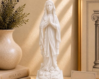 May include: A white marble-look statue of a figure with hands clasped in prayer, wearing a draped robe and a veil, holding a rosary. The figure stands on a rectangular base decorated with sculpted roses. A small card reads estatuesshop Greek statues.