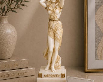 以下が含まれることがあります： 前面にギリシャ語のテキスト「ΑΦΡΟΔΙΤΗ」が刻まれた長方形の台座の上に立つ、クリーム色の女神アフロディーテの像。長い流れるような髪、ゴールドトーンの腕輪、腰にドレープ状の衣服をまとった姿が特徴です。