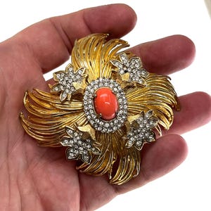 May include: A gold-toned brooch with a floral design. The center features an oval, coral-colored stone surrounded by a ring of clear crystals. Four smaller, crystal-studded flower accents are placed around the center. The brooch has a textured, layered design.