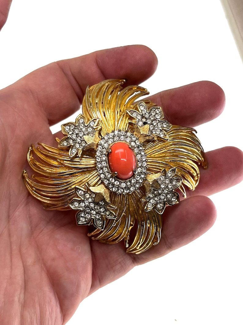 May include: A gold-toned brooch with a floral design. The center features an oval, coral-colored stone surrounded by a ring of clear crystals. Four smaller, crystal-studded flower accents are placed around the center. The brooch has a textured, layered design.