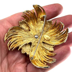 May include: A gold-toned, vintage brooch in a floral design. The brooch features layered, textured petals radiating from a central point. A pin and clasp are visible on the back. The brooch is held in a hand.
