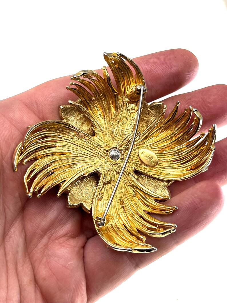 May include: A gold-toned, vintage brooch in a floral design. The brooch features layered, textured petals radiating from a central point. A pin and clasp are visible on the back. The brooch is held in a hand.