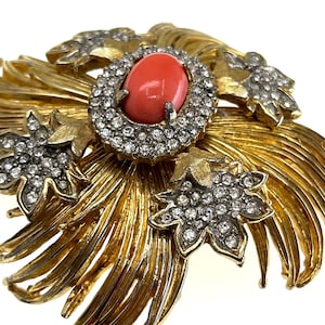 May include: A gold-toned brooch with a central oval coral-colored stone encircled by clear crystals. The brooch features stylized gold leaves and additional crystal-studded floral accents, creating a vintage, ornate design.