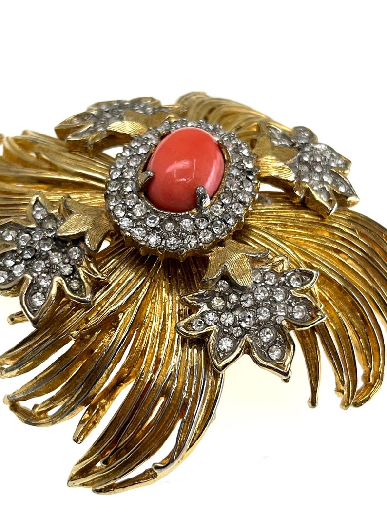 May include: A gold-toned brooch with a central oval coral-colored stone encircled by clear crystals. The brooch features stylized gold leaves and additional crystal-studded floral accents, creating a vintage, ornate design.