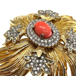 May include: A gold-toned brooch with a central oval coral-colored stone surrounded by clear crystals. The brooch features a floral design with additional crystal accents and a textured, radiating pattern.