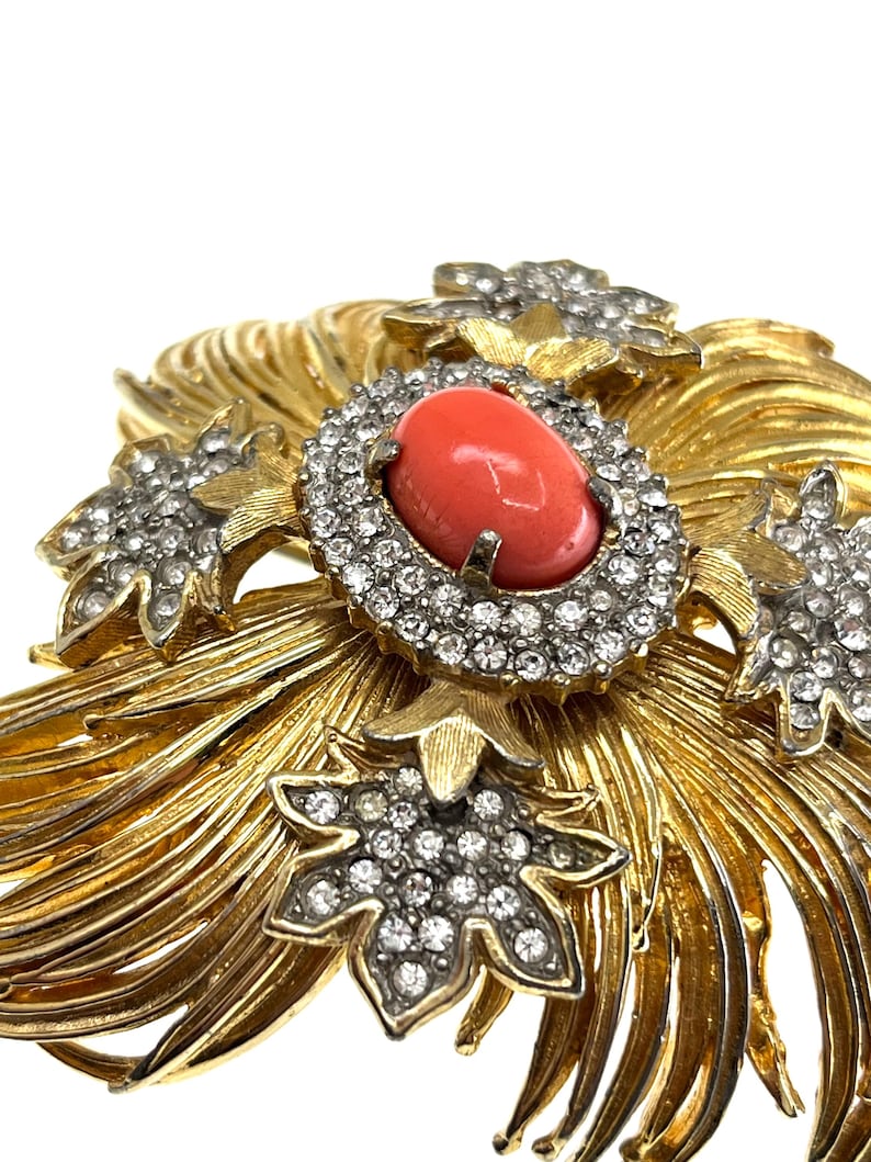 May include: A gold-toned brooch with a central oval coral-colored stone surrounded by clear crystals. The brooch features a floral design with additional crystal accents and a textured, radiating pattern.