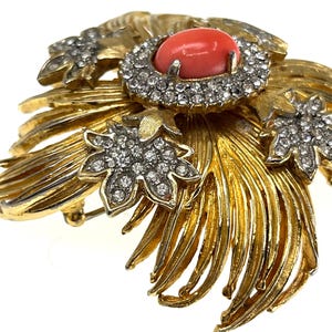 May include: A gold-toned brooch with a central coral-colored stone surrounded by clear crystals. The brooch features a floral design with detailed, textured leaves and additional crystal accents, creating a vintage aesthetic.