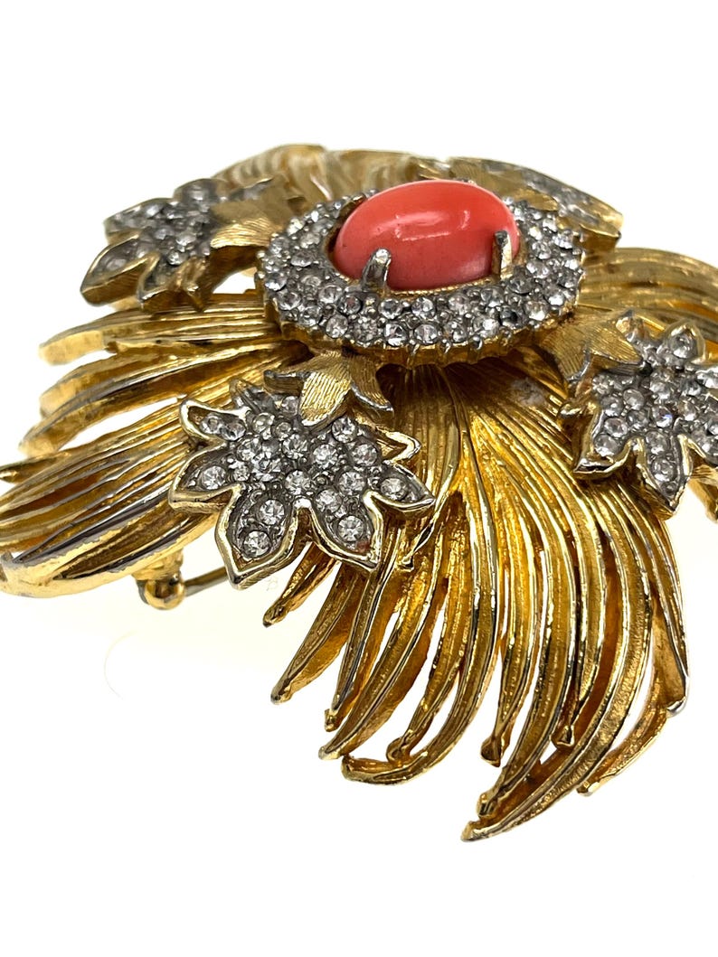 May include: A gold-toned brooch with a central coral-colored stone surrounded by clear crystals. The brooch features a floral design with detailed, textured leaves and additional crystal accents, creating a vintage aesthetic.