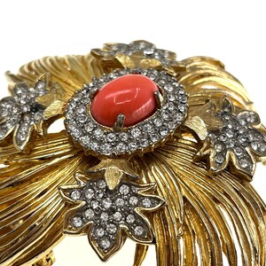 May include: A gold-toned brooch with a central oval coral-colored stone surrounded by a ring of clear crystals. The brooch features a floral design with additional crystal accents and a textured, feathery pattern.