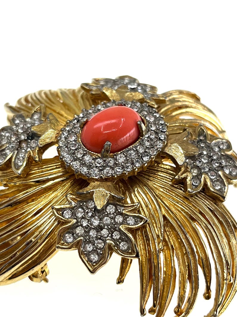 May include: A gold-toned brooch with a central oval coral-colored stone surrounded by a ring of clear crystals. The brooch features a floral design with additional crystal accents and a textured, feathery pattern.