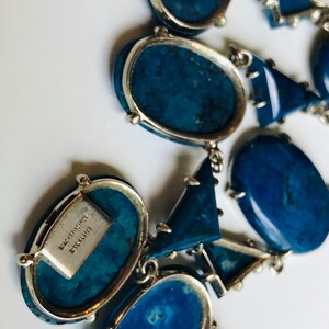 May include: A blue and silver statement necklace with a variety of geometric shapes. The necklace is made of metal and features a signature tag that reads "Baccarat Signature".