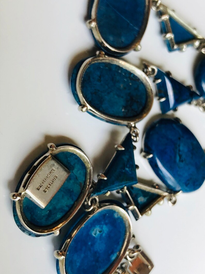 May include: A blue and silver statement necklace with a variety of geometric shapes. The necklace is made of metal and features a signature tag that reads "Baccarat Signature".