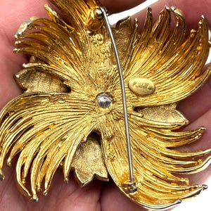 May include: A gold-toned metal brooch in a floral design. The brooch has a pin on the back for attachment. The design features layered petals and a textured finish. The brooch is likely vintage.